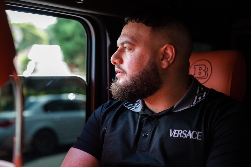 A man looks out the window of a car. The car's interior is orange. The man is wearing a shirt with 'Versace' on the chest.
