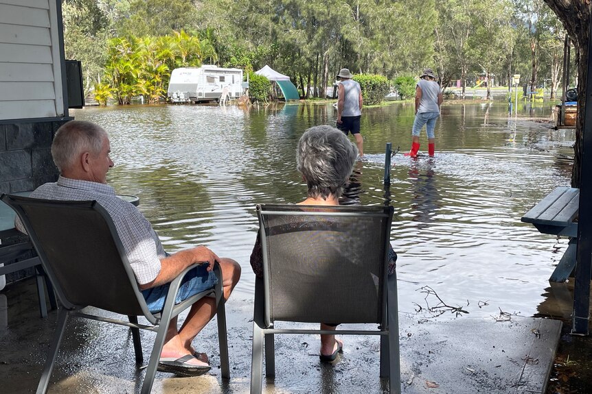 A man and woman sit in chairs looking out over floodwater in a caravan park.