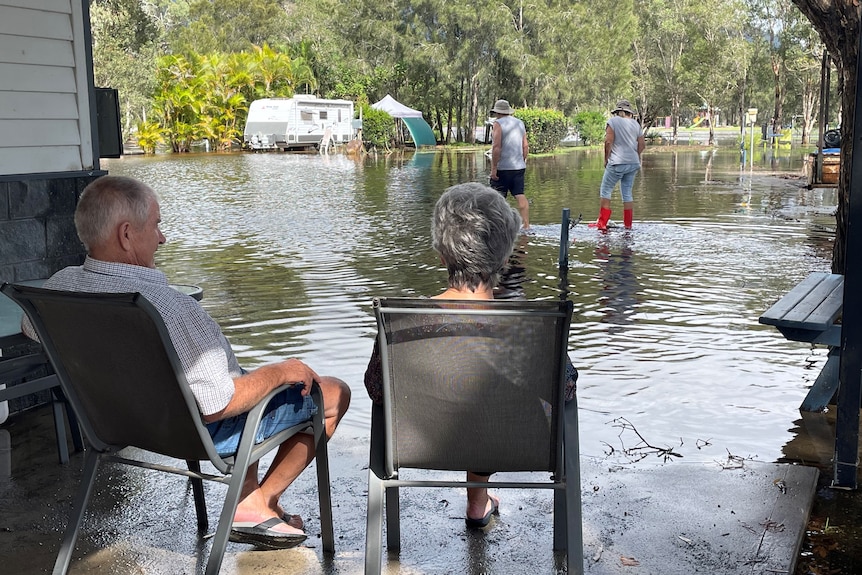 A man and woman sit in chairs looking out over floodwater in a caravan park.