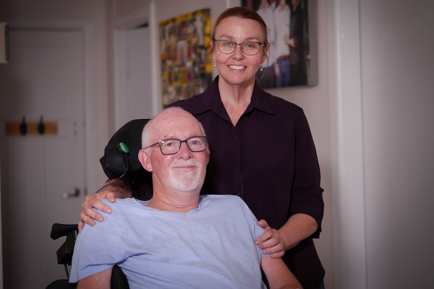 A man in a wheelchair with his wife standing behind him and a family portrait on the wall behind them.