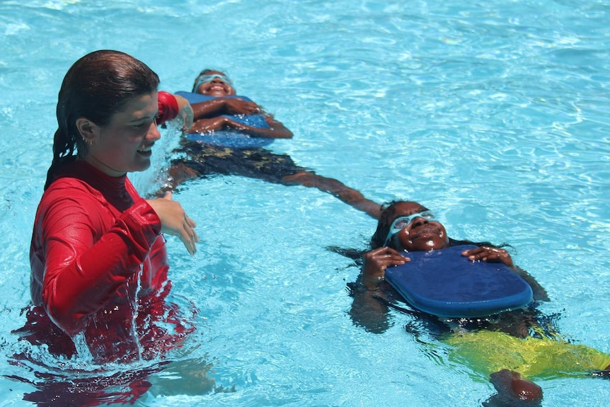 An adult swim instructor assists as two young children practice floating on their backs in clear water