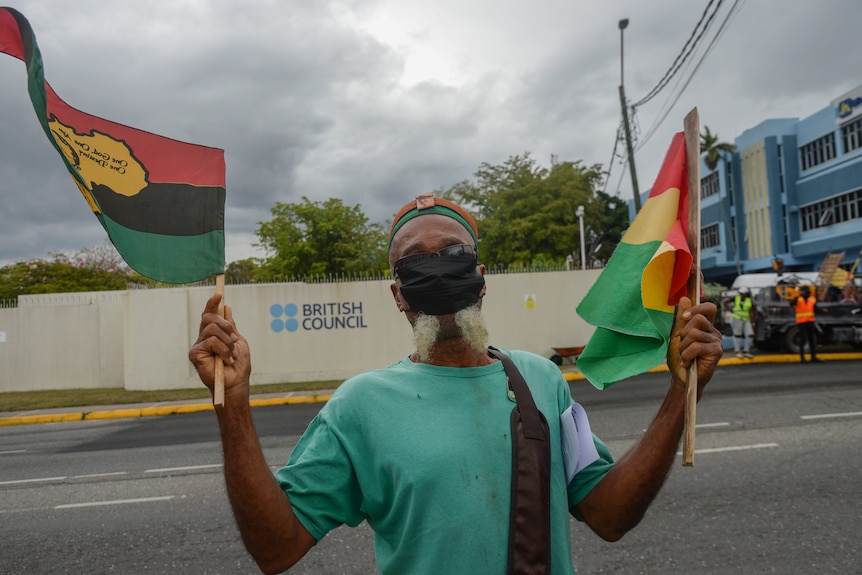 a man with a jamaican flag in each hand, wearing a green t shirt and a black mask