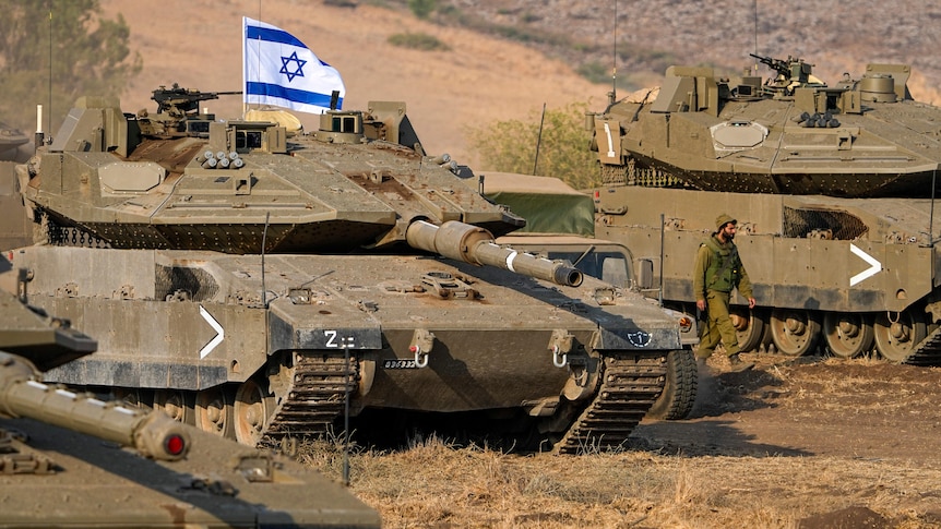 Three tanks, one flying the Israeli flag, sit in position on brown grass as a hemleted soldier walks between them.