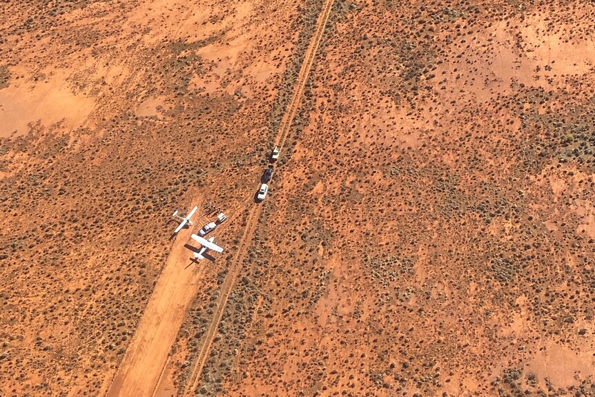 Aerial photo of two planes on a red dirt airstrip, with a car and a trailer beside them, three cars on a dirt road nearby.