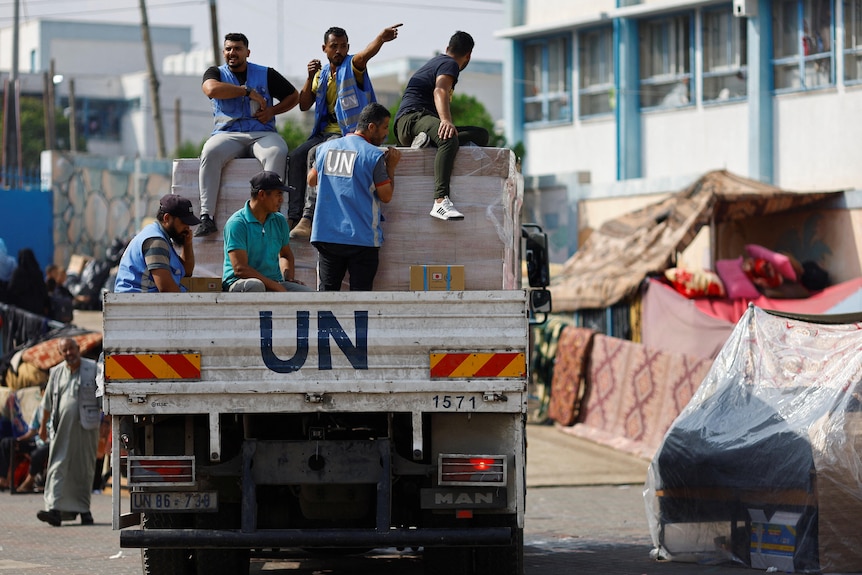A truck is driving with the letters UN, there are boxes in the back with people sitting on top of the boxes wearing UN vests.
