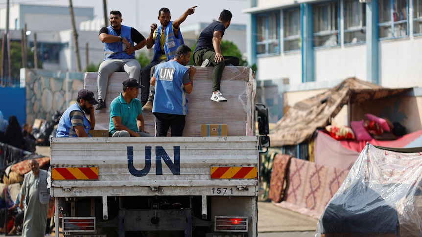 A truck is driving with the letters UN, there are boxes in the back with people sitting on top of the boxes wearing UN vests.