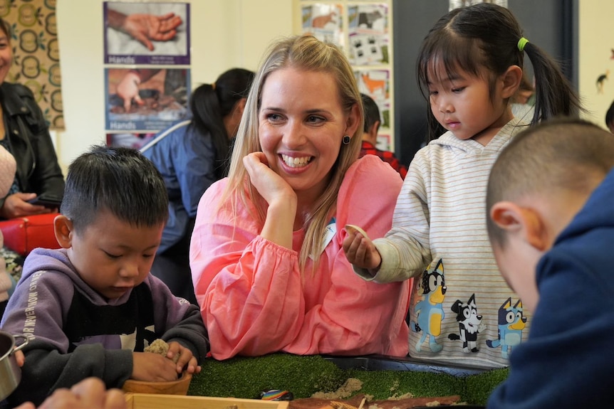 A smiling woman rests her arm on a table surrounded by young children playing