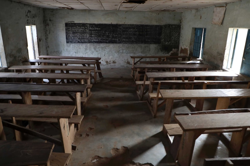 An empty classroom with wooden tables and bench seats and damaged floor, walls and ceiling.