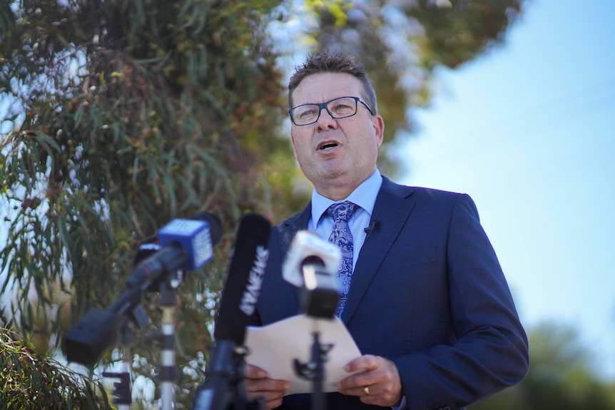 A man wearing a suit and tie and glasses reads from a piece of paper standing behind microphones