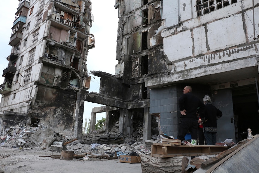 Persone in piedi accanto a un grande condominio che è stato danneggiato durante pesanti combattimenti e bombardamenti.