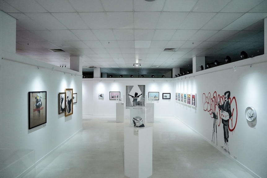 A large room with white walls and shiny white tiles showcases several Banksy artworks.