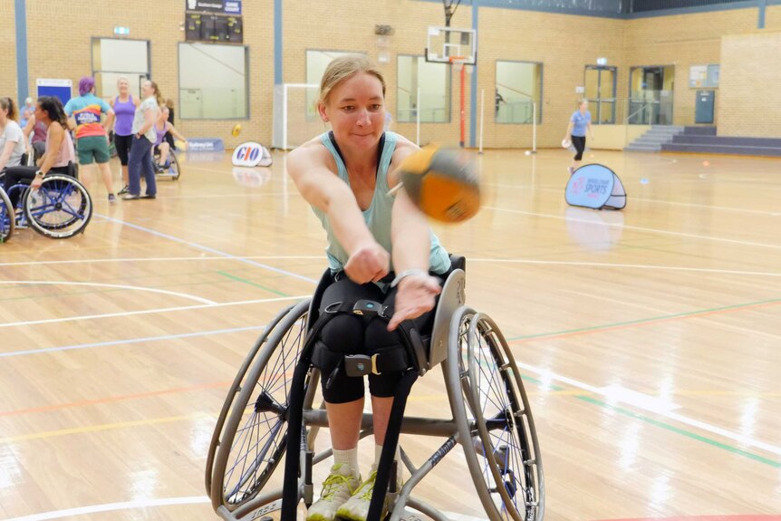 A woman handballs a football from a wheelchair while on a basketball court.