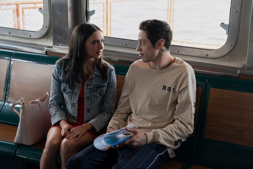 A young woman in red dress and denim jacket and young man in long sleeve t-shirt sit looking at each other in ferry cabin.