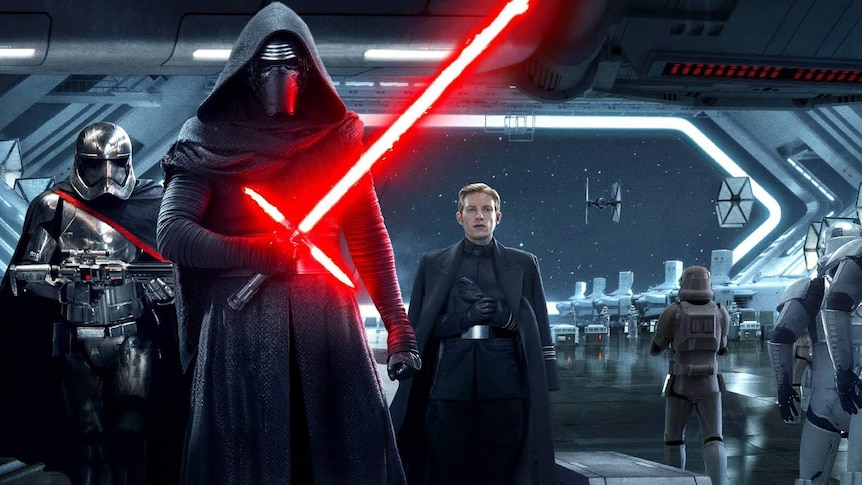 Kylo Ren dressed in black holds a red lightsabre while standing in a spaceship, flanked by two henchman.