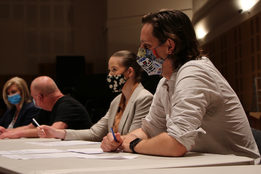 People wearing face masks sit behind a table with notepads and pens as they watch someone perform