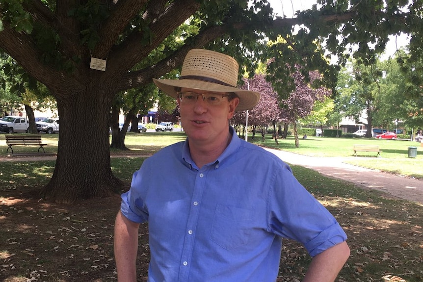 Man wearing hat and blue shirt stands with hands on hips under a tree.
