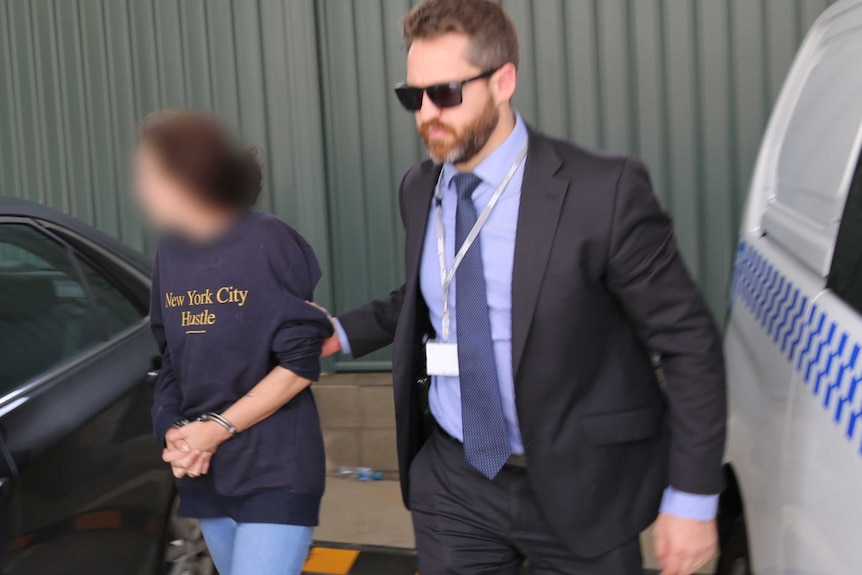 A handcuffed woman whose face is blurred is led through a car park by police.