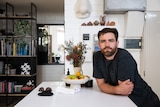 Alex Goad looks into the camera as he leans against a white kitchen counter carrying a fruit bowl, and a flower vase.
