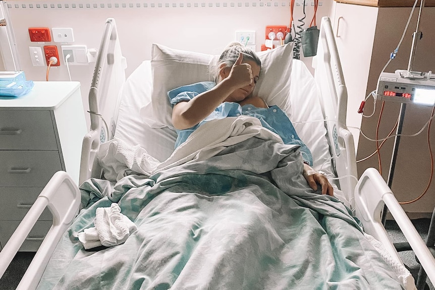 A woman in a hospital bed looking away from the camera with her thumbs up