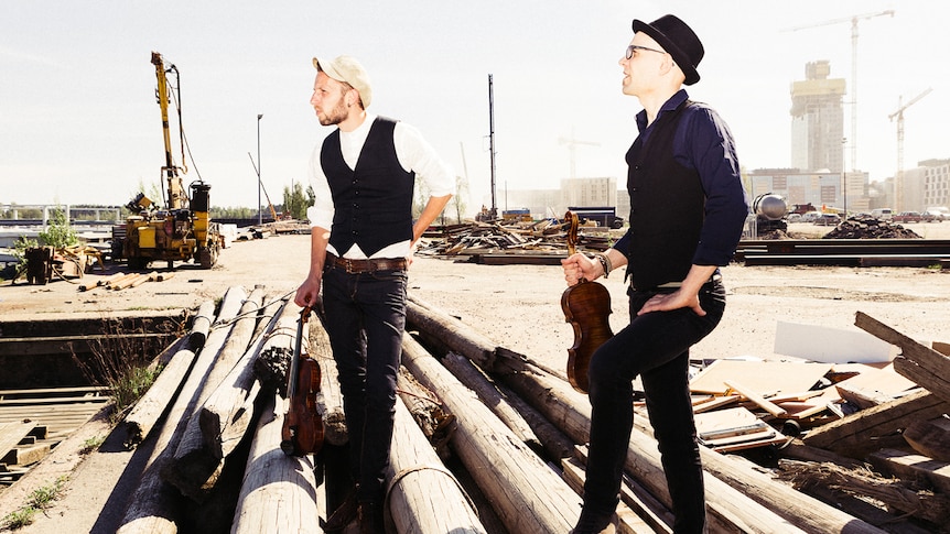 Esko and Tero pose in a lumberyard, standing on sawn bits of wood. They hold their violins and look into the distance.