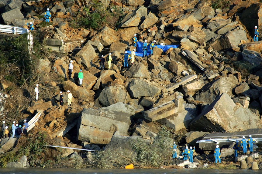 Rescue workers spread a blue tarp across a portion of collapsed cement and rock strewn across a road