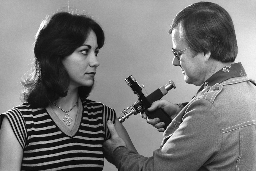 A black and white photo of a woman receiving an injection via jet injector from a man.