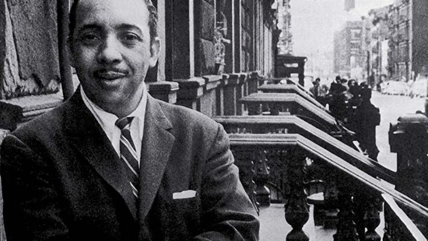 A monochrome photo of Red Garland standing on a street in Harlem with his suit on, looking directly at the camera.