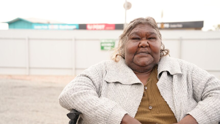 A photo showing an Indigenous woman sitting in a chair outside with building in the background.