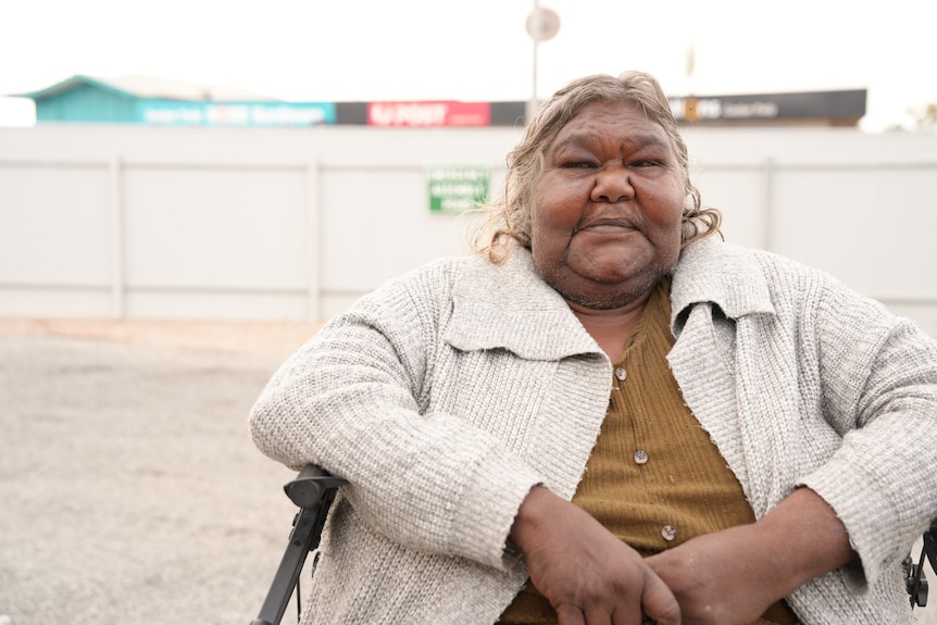 A photo showing an Indigenous woman sitting in a chair outside with building in the background.