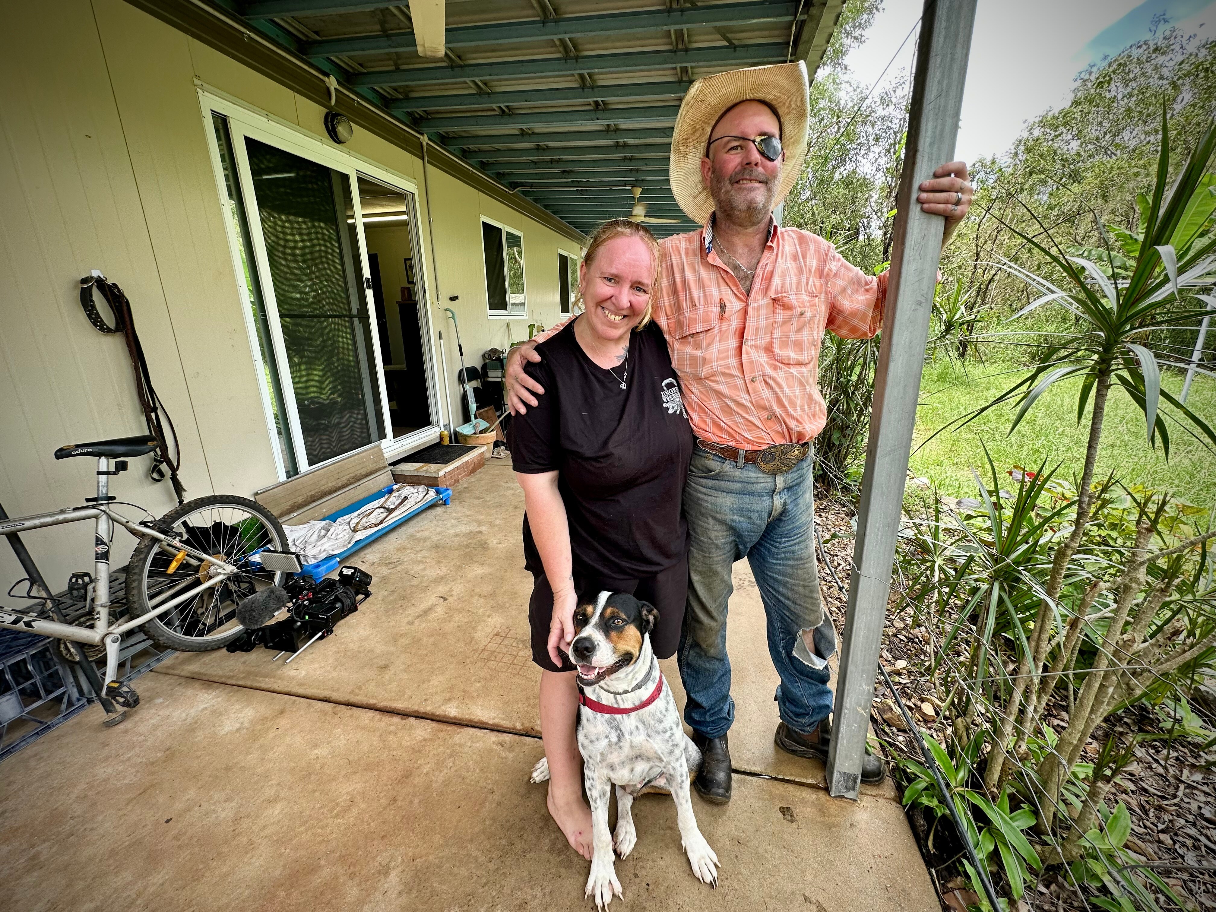 A woman wearing a black t-shirt, patting her dog standing with her husband who's wearing jeans, a straw hat, and eye patch