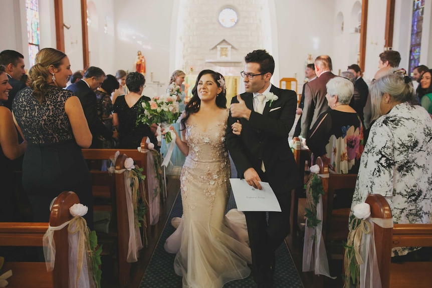 Nas and Tom walk up the aisle after their wedding ceremony, smiling and among friends and family