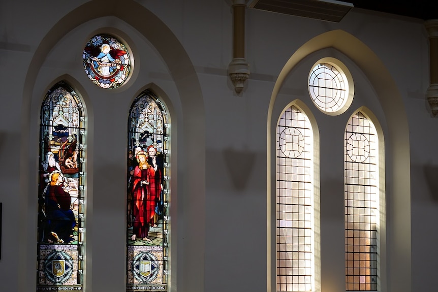 A stained glass window with religious images next to a plain lead light window, photographed from inside a church