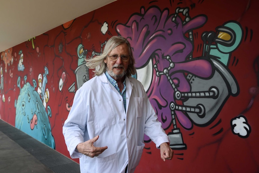 A man in a white coat walks in front of a graffitied wall.