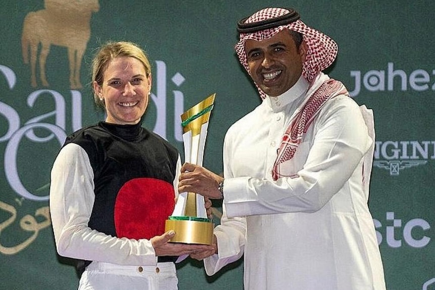 A woman standing holding  trophy alongside a man wearing a traditional Saudi thobe.