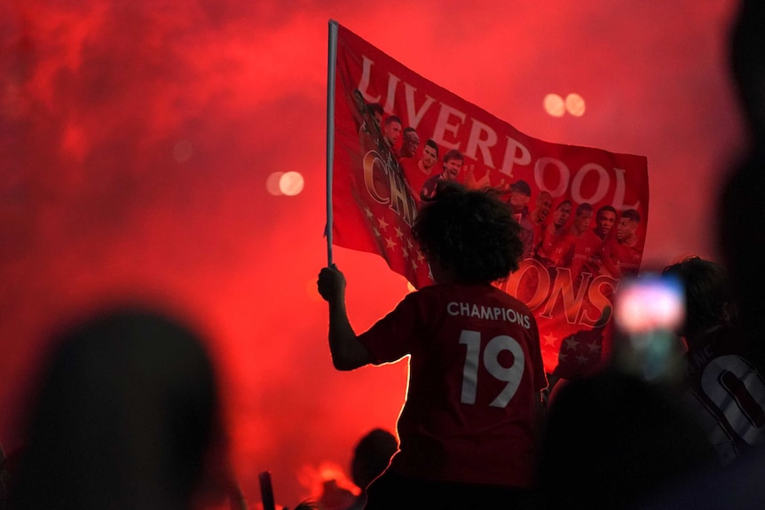 A Liverpool fan is seen through red smoke, waving a banner while wearing a "Champions 19" shirt.