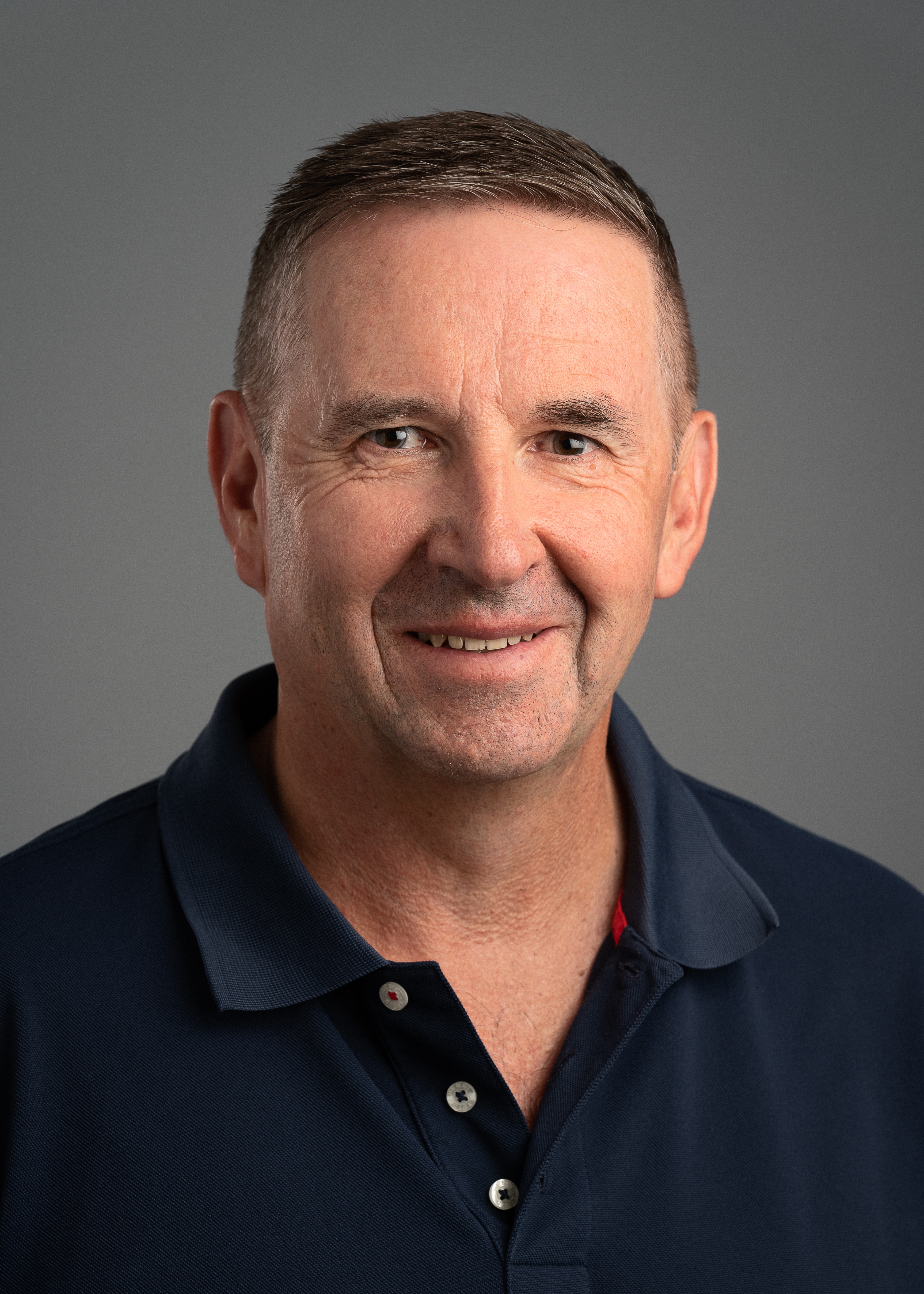 Um homem de meia idade com uma camisa pólo preta simples olha para a câmera, o fundo é cinza. 