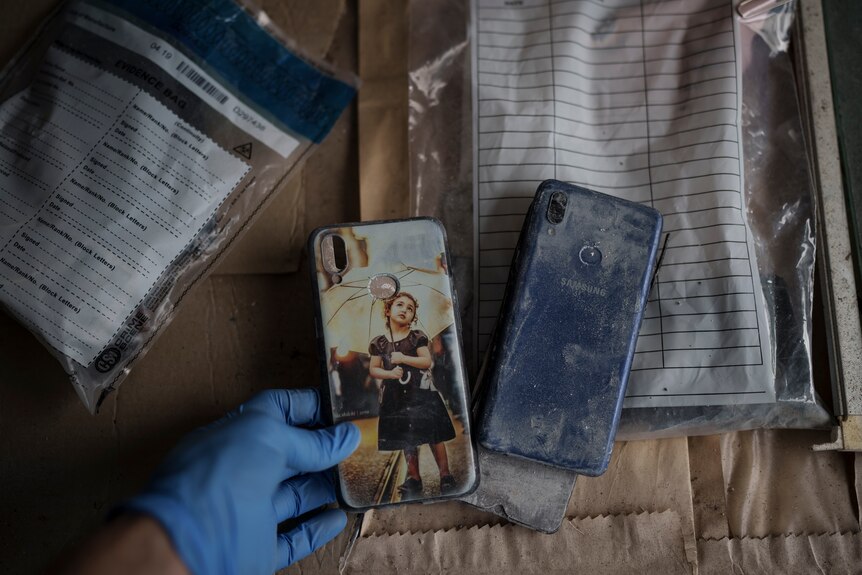 A blue latex glove is pictured holding a dusty smartphone, another one lying next to it. Behind it are evidence bags.