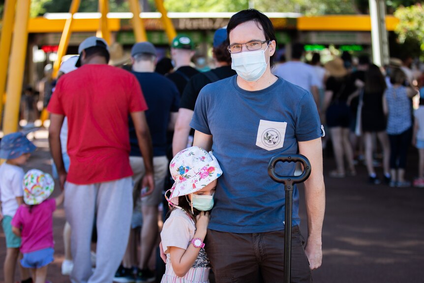 A man with a mask stands outside the zoo, while a girl hugs his side closely.