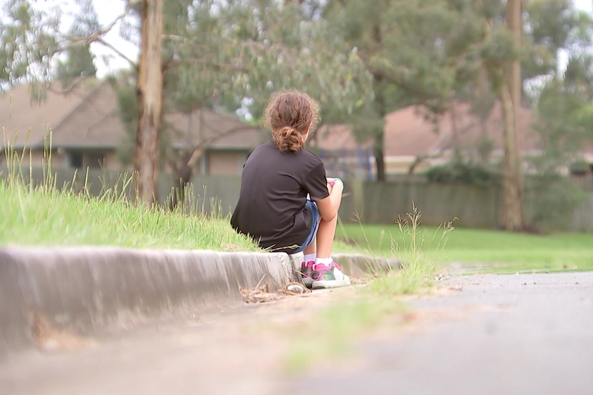 Young girls sits on a curb with knees huddled up, back turned to camera.