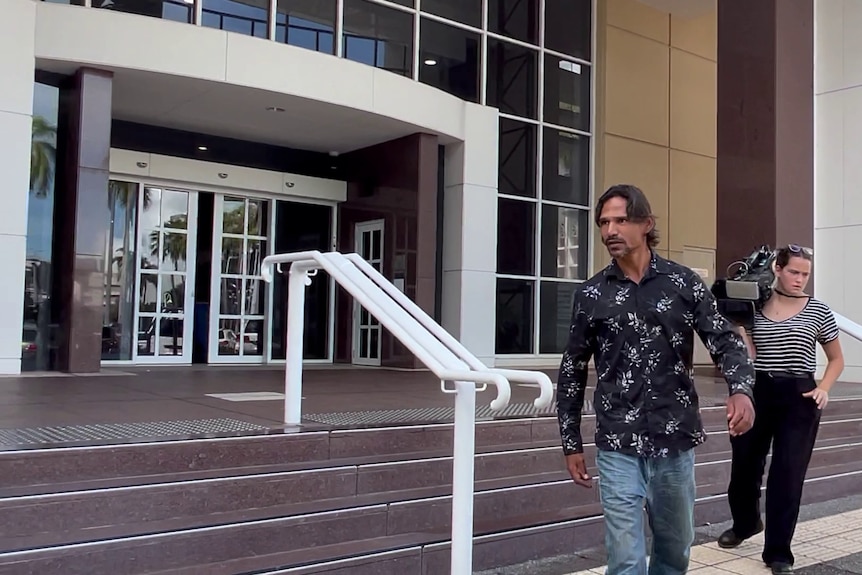 Un hombre aborigen con una camisa negra con flores blancas afuera de las puertas delanteras de la Corte Suprema de Darwin.