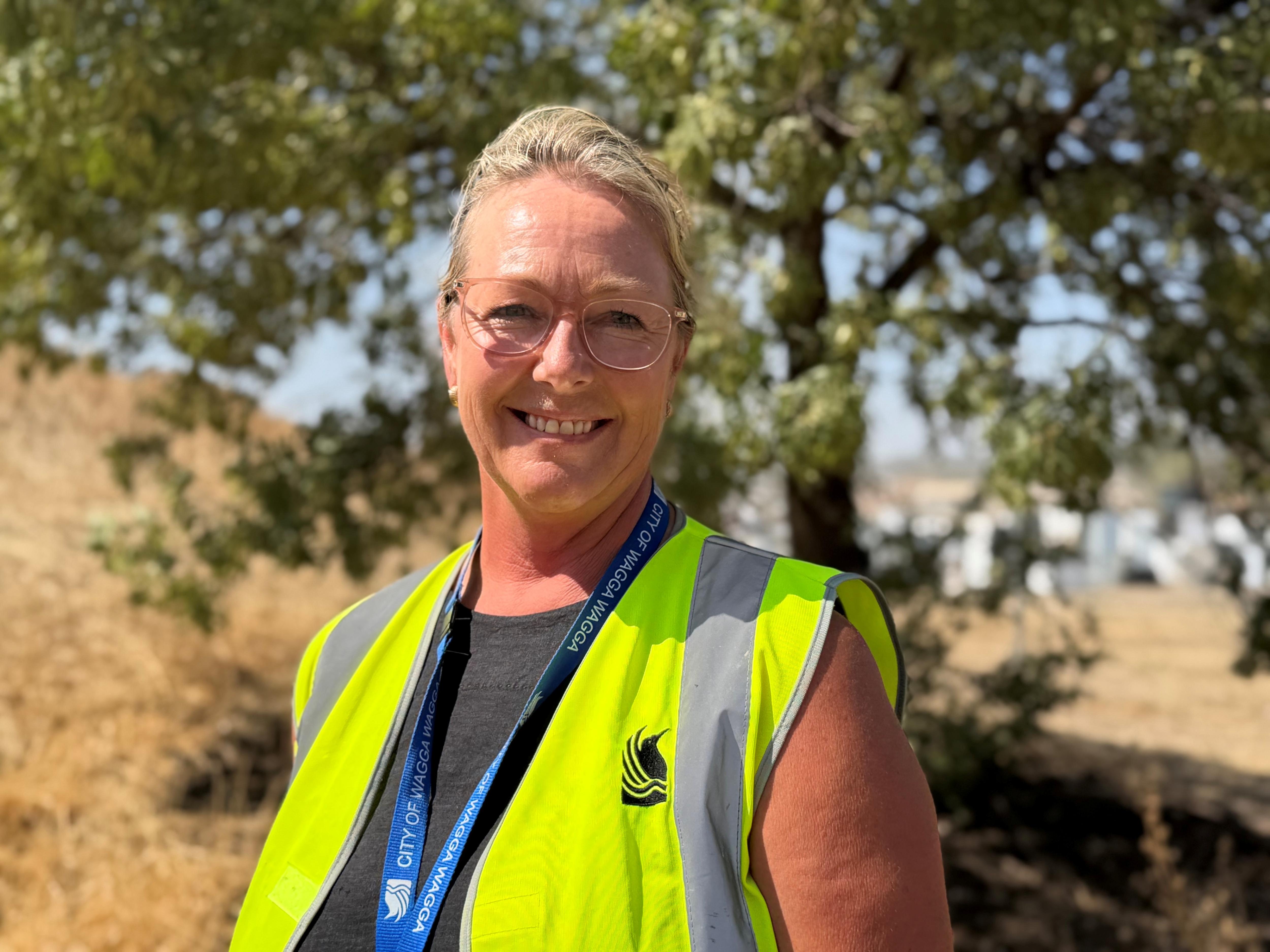 Uma mulher loira de óculos está com um colete de alta visibilidade em frente a uma árvore.