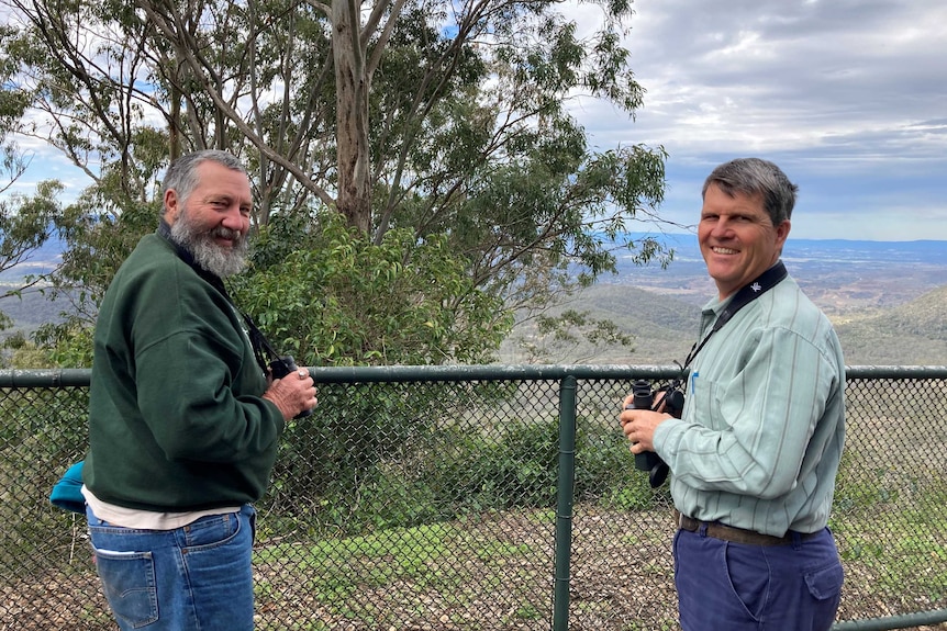 Two men with binoculars standing on a hill overlooking bushland and smiling.