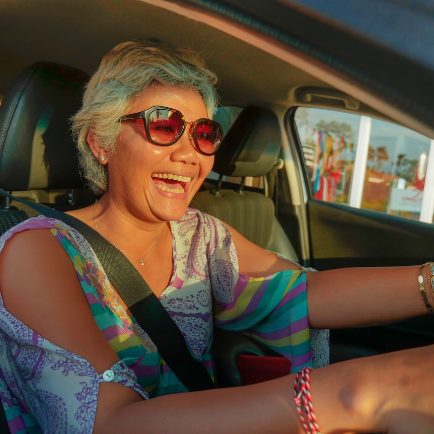 A woman smiles, wears sunglasses and a colourful, summer-style top, while sitting behind the wheel of a car.