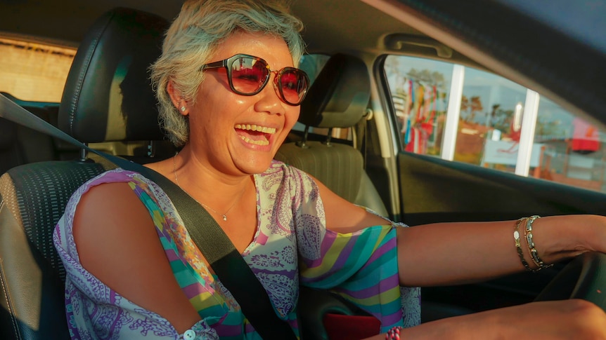 A woman smiles, wears sunglasses and a colourful, summer-style top, while sitting behind the wheel of a car.