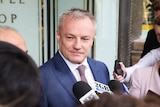 A middle-aged man in a blue suit speaks to media, holding microphones, at a scrum outside court.