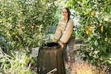 Koren Helbig smiles while standing next to a compost bin in a sunny, lush garden.