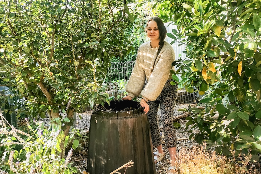 Koren Helbig smiles while standing next to a compost bin in a sunny, lush garden.
