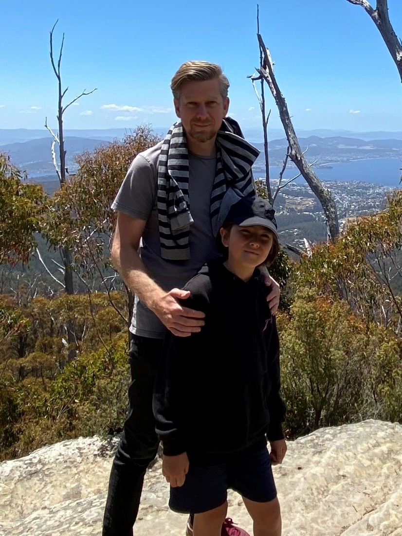 A smiling man stands with his young son at a scenic viewpoint, with mountains and blue sky in the background.