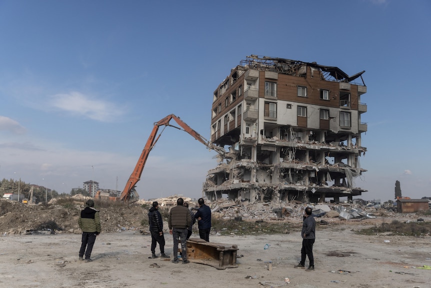 La gente guarda un veicolo da costruzione smantellare un edificio distrutto in Turchia.
