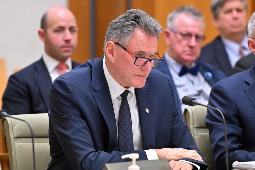 Ross McEwan in a suit and glasses sits at a desk in Parliament House with men sitting behind him